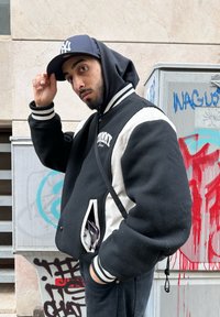 Čierna a krémová varsity bunda so pruhovanými manžetami, prednými vreckami a kapucňou. Doplnky zahŕňajú čiernu baseballovú čiapku. Mestské pozadie.