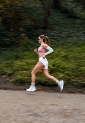 Atleta che corre su un sentiero, indossando un reggiseno sportivo rosa, una manica verde chiaro, pantaloni corti bianchi e scarpe da corsa bianche.