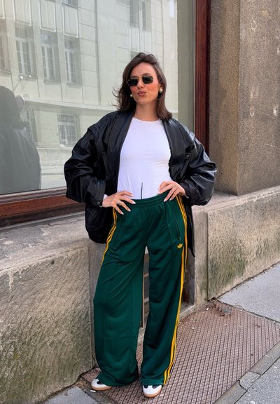 Chaqueta de cuero negra de gran tamaño, camisa blanca ajustada, pantalones de chándal verdes con rayas amarillas y zapatillas blancas. Pendientes circulares. Fondo urbano.