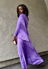 Abito viola fluido con maniche ampie e scollatura alta, caratterizzato da una trama liscia. Completato da una pochette dorata e tacchi rosa.