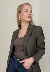 Blazer marron sur mesure porté sur un débardeur marron côtelé, associé à un jean bleu foncé. Le mannequin porte de grandes boucles d'oreilles en or. Textures lisses visibles.