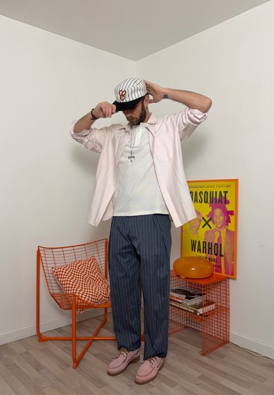 Hombre ajustando gorra a rayas, vistiendo camisa rosa claro, camiseta blanca, pantalones de rayas finas, de pie junto a una silla de alambre naranja y mesa auxiliar a juego con un póster de arte.