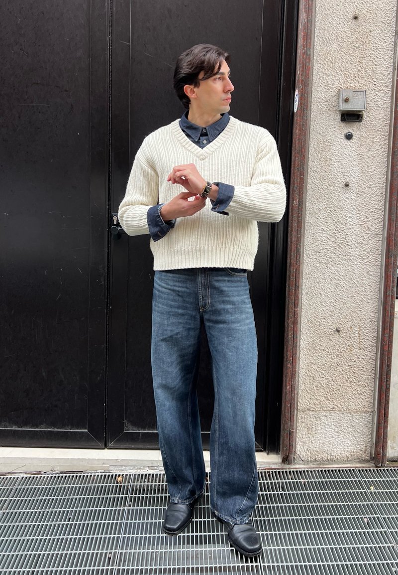 Homme portant un pull en maille blanc, une chemise bleue, un jean à jambes larges et des chaussures noires, debout en train de régler sa montre devant une porte noire et un mur gris.