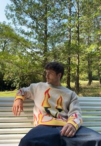 Γκρι πουλόβερ με κόκκινες και κίτρινες φλόγες, ριγωτές μανσέτες και χαλαρή εφαρμογή. Μπλε τζιν παντελόνι, καθισμένος σε ένα λευκό παγκάκι σε μια καταπράσινη περιοχή.