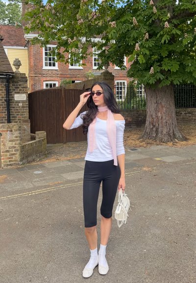 Mujer con gafas de sol, top blanco de hombros descubiertos, bufanda rosa, pantalones capri negros y zapatos blancos, está de pie en la calle cerca de una casa de ladrillos y un árbol grande.