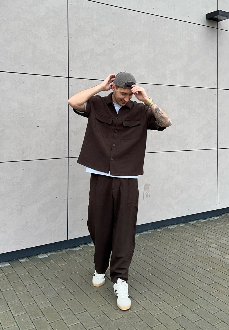 Brun skjorta med knappar fram och två bröstfickor, kombinerad med matchande byxor med lös passform och vita sneakers på ett kaklat underlag. Grå keps på huvudet.