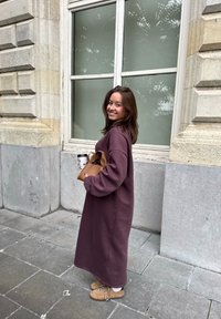 Lang, losvallende paarse sweatshirt jurk met lange mouwen, gecombineerd met lichtbruine laarzen en een bruine handtas. Houdt een koffiekop vast.