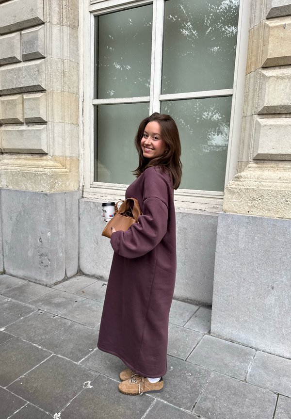 Flowing, oversized purple sweatshirt dress with long sleeves, teamed with light brown boots and a brown handbag. Holding a cup of coffee.