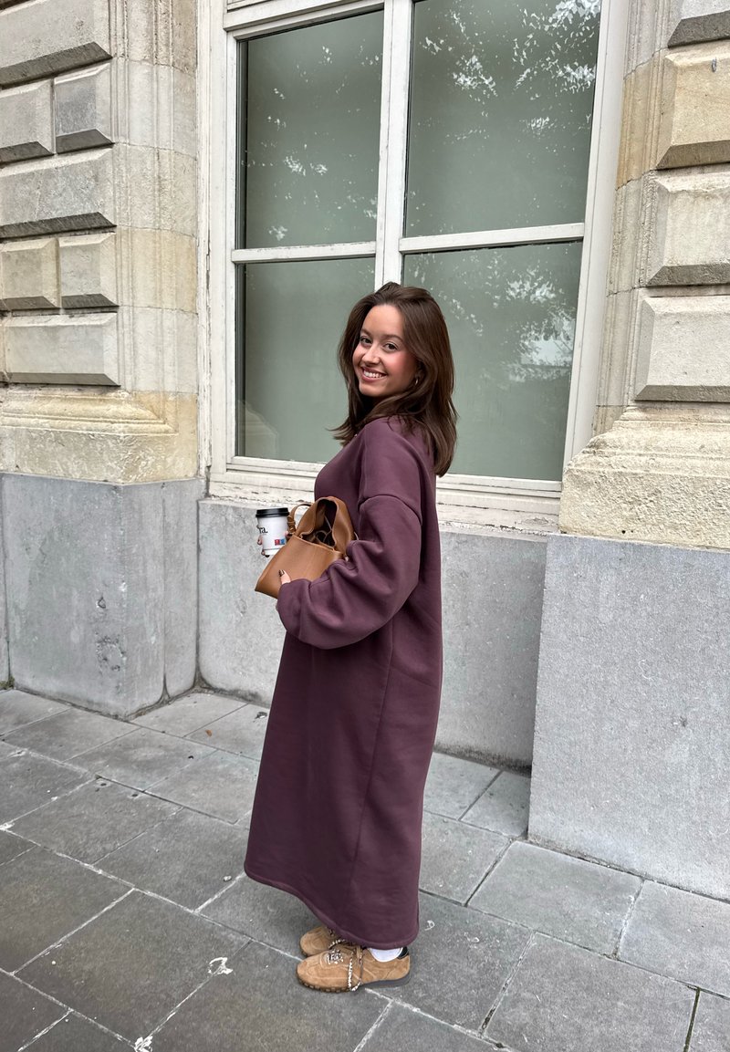 Abito felpato viola lungo e largo con maniche lunghe, abbinato a stivali marrone chiaro e una borsa marrone. Tiene una tazza di caffè.