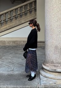 Zwarte cropped sweater, witte shirt eronder, sheer geruite rok in blauwtinten, zwarte loafers, kleine zwarte handtas, staande naast een stenen kolom.