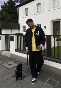 Černá varsity bunda s bílými pruhy a písmenem "J", pod ní žluté tričko, volné černé kalhoty a malý pes na vodítku.