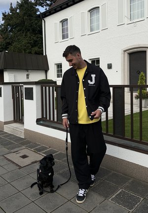 Chaqueta varsity negra con rayas blancas y letra "J", camiseta amarilla debajo, pantalones negros holgados y un perro pequeño con correa.