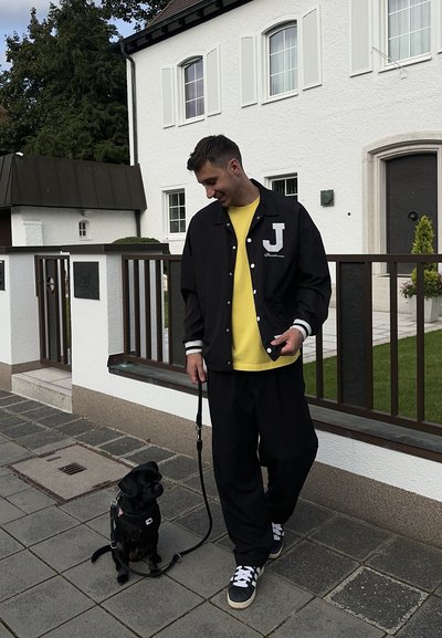 Chaqueta varsity negra con rayas blancas y letra "J", camiseta amarilla debajo, pantalones negros holgados y un perro pequeño con correa.
