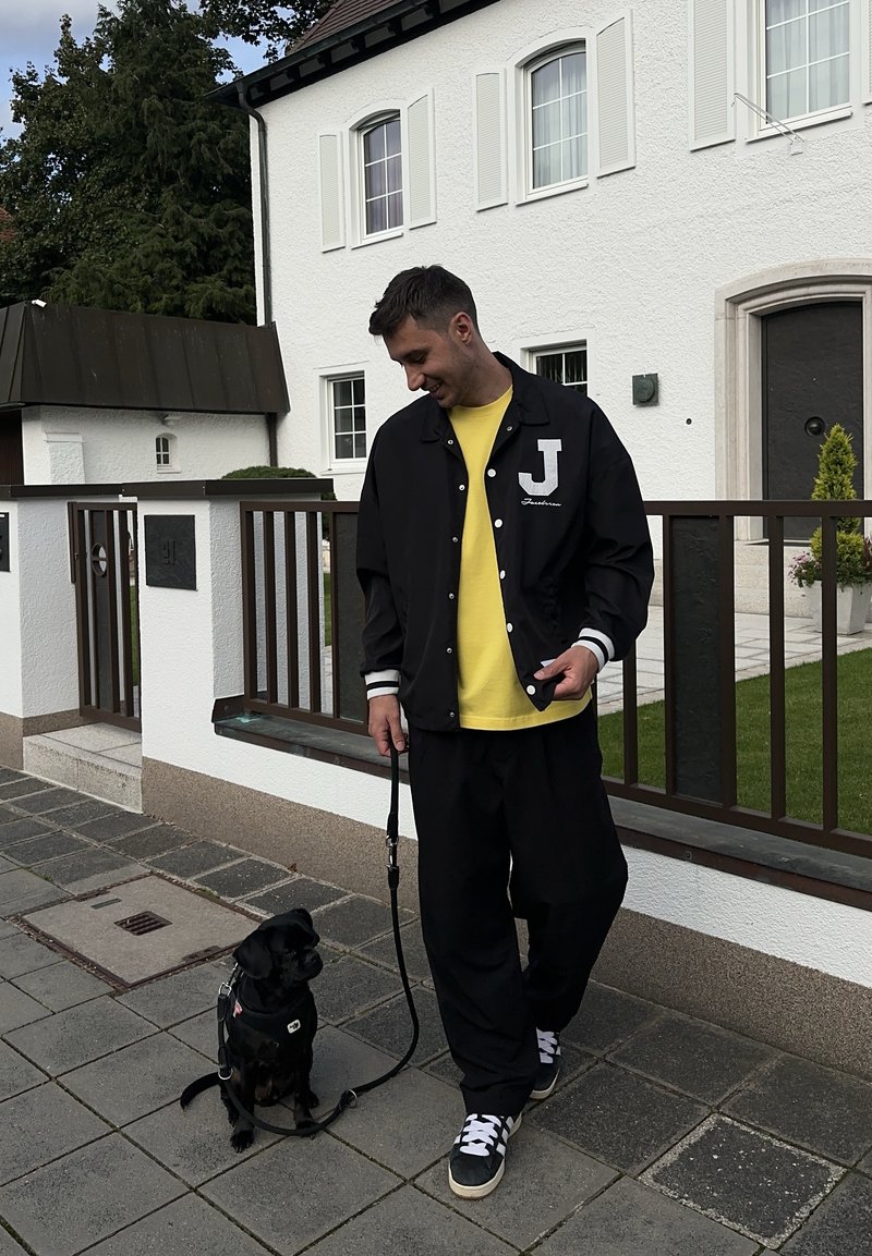 Veste de varsity noire avec des rayures blanches et la lettre "J", chemise jaune en dessous, pantalon noir ample, et un petit chien en laisse.