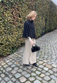 Top nero in stile mantello con colletto alto, pantaloni beige a gamba larga, borsa nera e stivali alla caviglia. Sentiero lastricato in pietra e sfondo di fogliame verde.