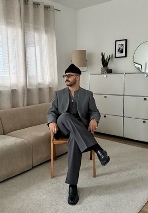 Hombre con traje gris y sombrero negro sentado en una silla de madera. La habitación cuenta con un sofá beige, cortinas claras, una planta en maceta y un espejo redondo.