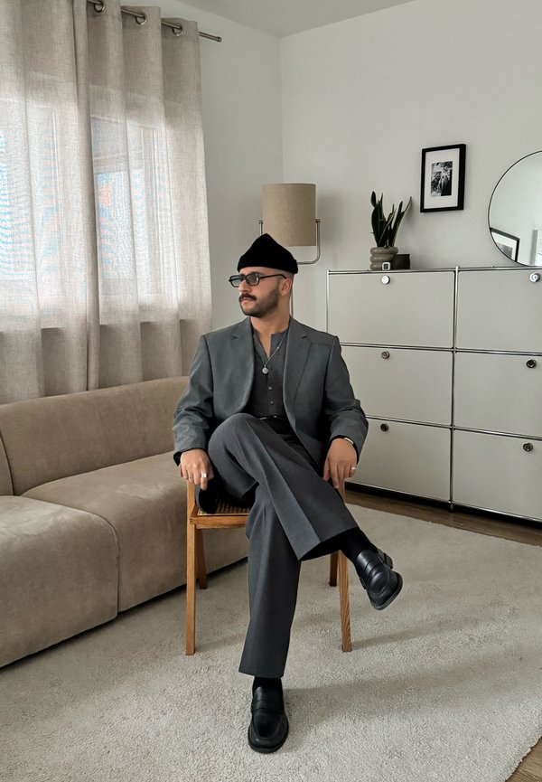 Man in een grijs pak en zwarte hoed zit op een houten stoel. De kamer heeft een beige bank, lichte gordijnen, een plantenpot en een ronde spiegel.