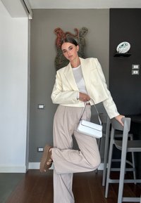 Witte cropped blazer over een wit topje, beige wijde broek en bruine sandalen. Houdt een kleine witte handtas vast, staand tegen een grijze muur.