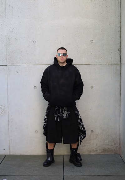 Sudadera negra de gran tamaño con efecto degradado, pantalones cortos negros, una camisa de cuadros atada a la cintura y botas de combate negras. Gafas de sol reflectantes puestas.