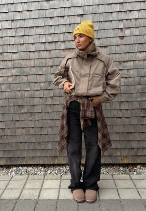 Veste beige courte avec un col montant, assortie à un jean foncé et des pantoufles brunes. Bonnet côtelé jaune et chemise à carreaux nouée à la taille.