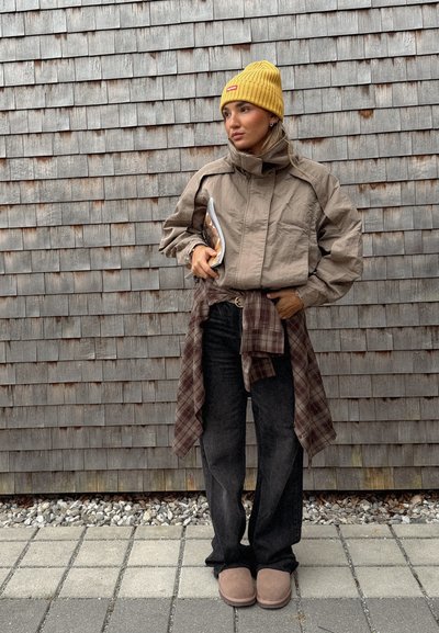 Chaqueta beige corta con un cuello alto, combinada con jeans oscuros y zapatillas color marrón. Gorro amarillo de canalé y una camisa de cuadros atada a la cintura.