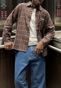 Brūna rūtainā krekls ar koka pogām pār baltu T-kreklu, kopā ar zilām džinsa biksēm. Fons ietver liftu paneļa detaļas.