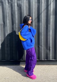 Um hoodie oversized azul brilhante, calças roxas de corte largo, tamancos cor de rosa e uma mala amarela com alça de corrente. Óculos de sol transparentes completam o visual.
