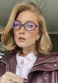 Stora, runda glasögon med lila ton och tunn mörk båge. En vinröd skinnjacka över en vit skjorta med rosa ränder och en slips.