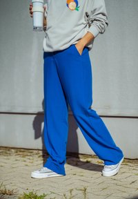 Mėlyni laisvi kelnės su lygaus paviršiaus, derinamos su baltais sportiniais bateliais. Pilkas megztinis su spalvinga vaisių grafika ir priekinė kišenė.