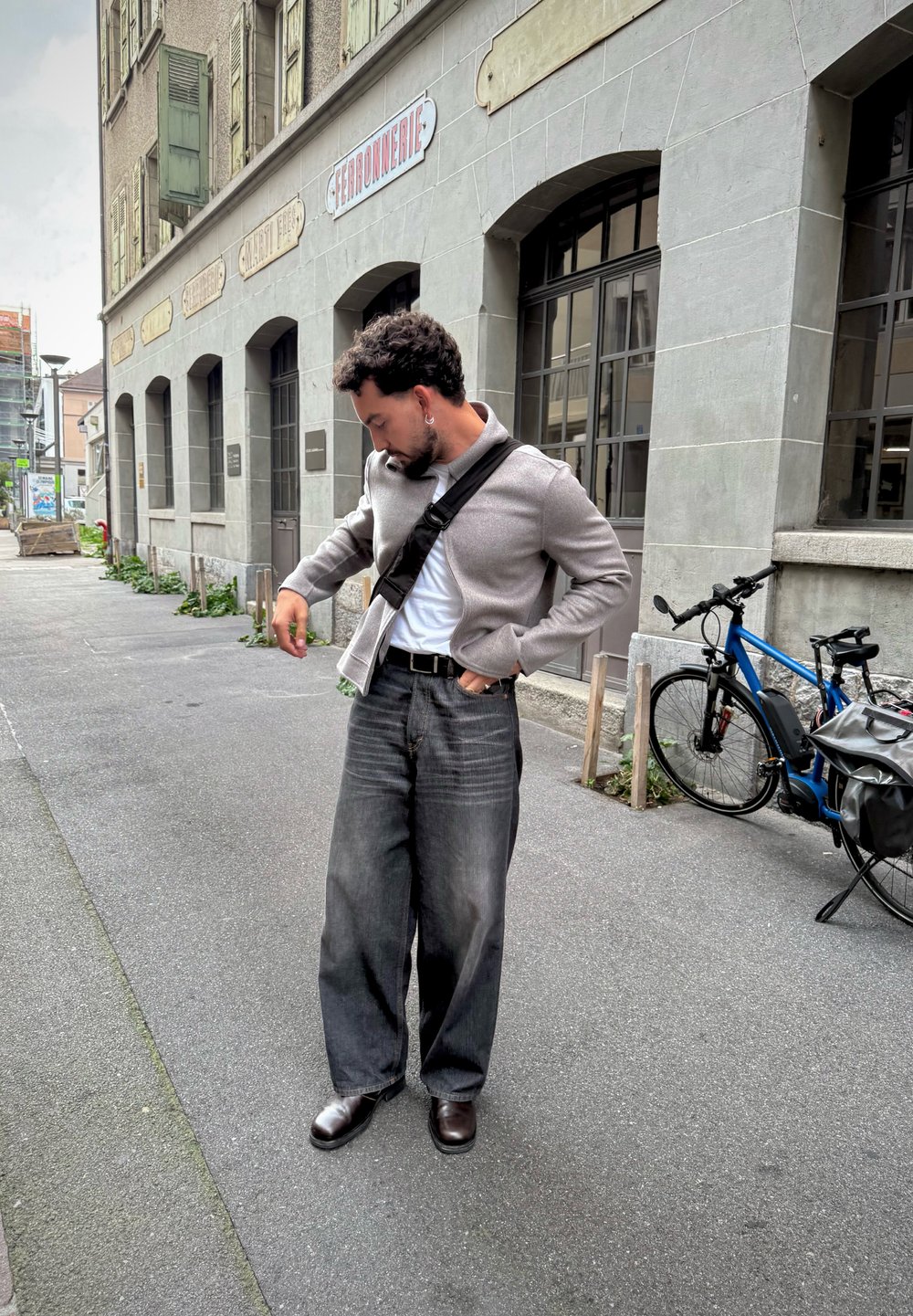 Férfi keresztpántos táskával igazítja a dzseki zsebéből a szélén, egy épület mellett, ahol íves ablakok találhatók, egy kék bicikli mellett, amely leparkolt.