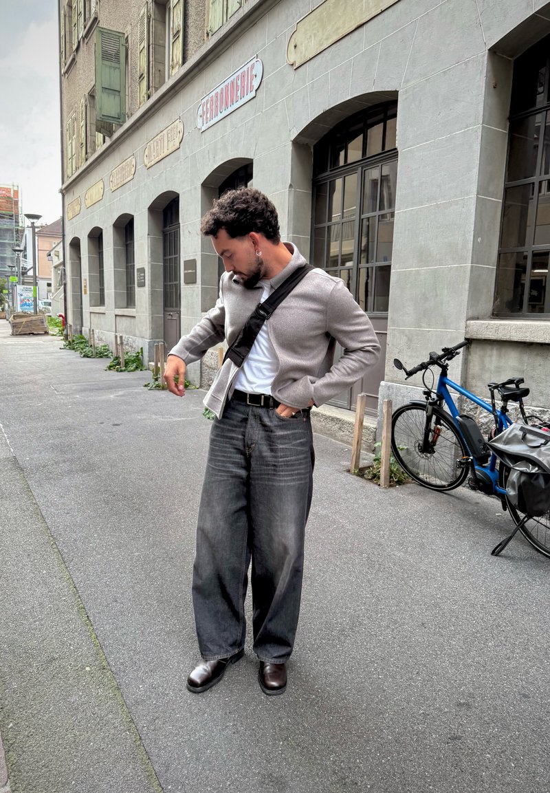Hombre con bandolera ajusta el bolsillo de la chaqueta en la acera junto a un edificio con ventanas arqueadas, al lado de una bicicleta azul estacionada.