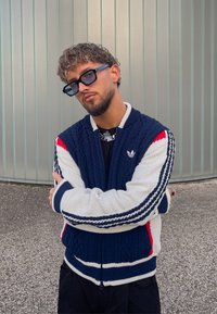 Cardigan tricotat bleumarin și alb, cu accente roșii, având manșete striate, închidere cu fermoar și un logo pe piept. Purtat peste un tricou negru.