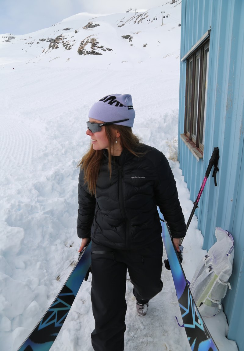 Femme en tenue d'hiver portant des skis, marchant à côté d'un bâtiment bleu dans un paysage montagneux enneigé.