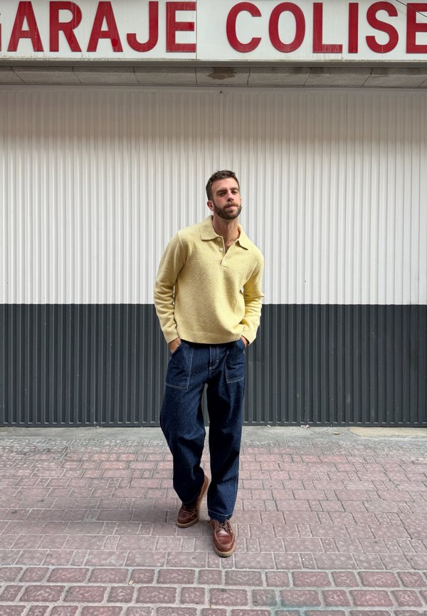 Hombre con barba que lleva un suéter amarillo, pantalones anchos azules y zapatos marrones, de pie sobre un pavimento de ladrillos frente a una pared ondulada en blanco y negro.