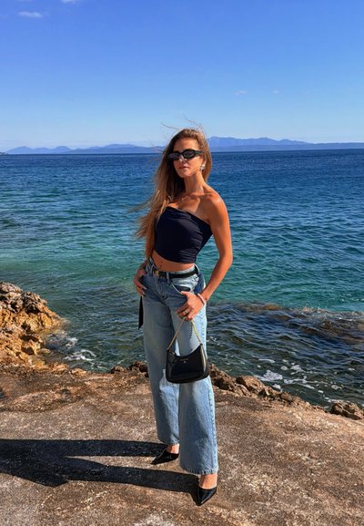 Mujer con cabello largo que lleva gafas de sol, top negro sin tirantes, vaqueros anchos, sosteniendo un bolso negro, de pie en la orilla rocosa junto al mar azul bajo un cielo despejado.