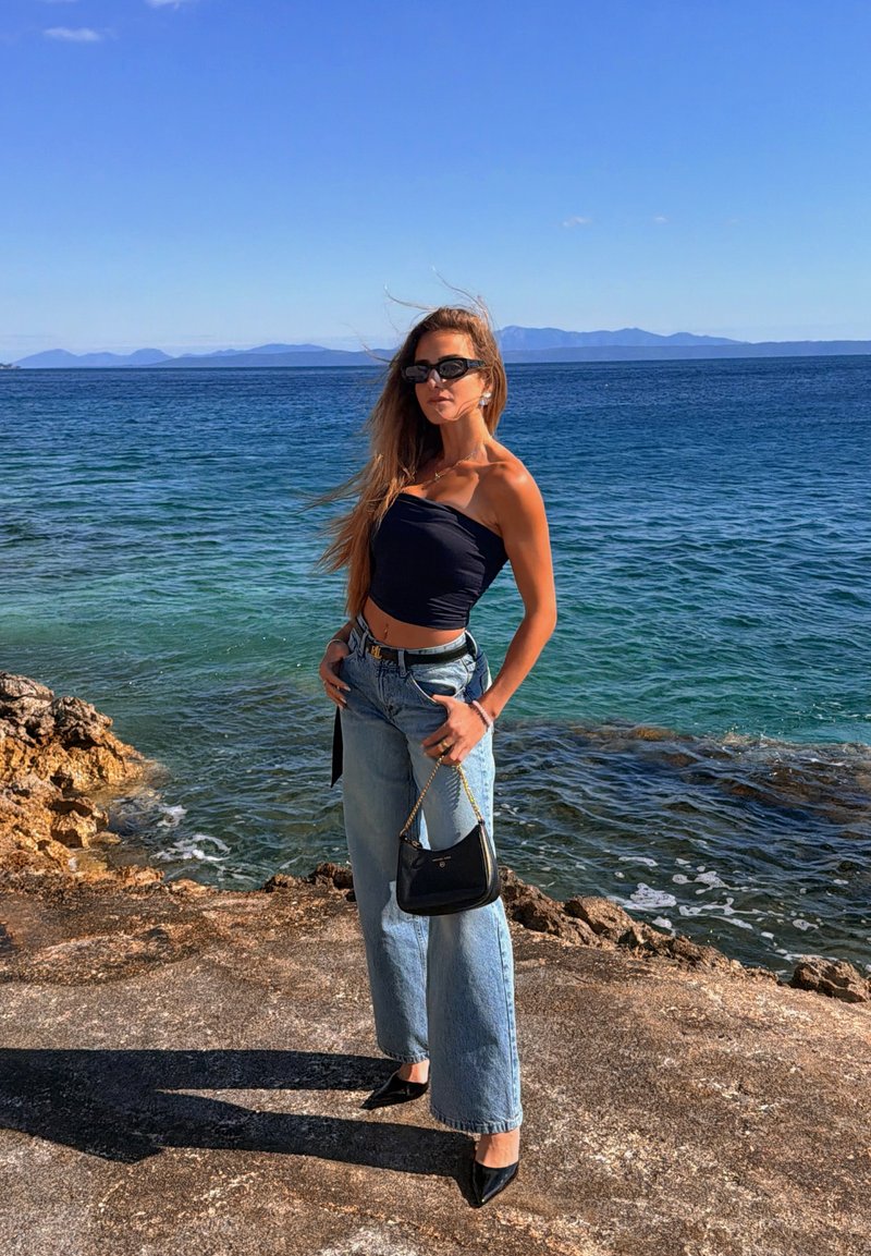 Mulher de cabelo comprido a usar óculos de sol, top preto, jeans largos, segurando uma mala preta, de pé numa costa rochosa junto ao mar azul sob céu limpo.