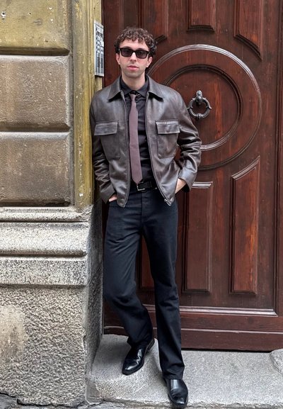 Chaqueta de cuero marrón con dos bolsillos en el pecho, camisa negra y corbata marrón. Pantalones y zapatos negros, apoyado contra una puerta de madera.