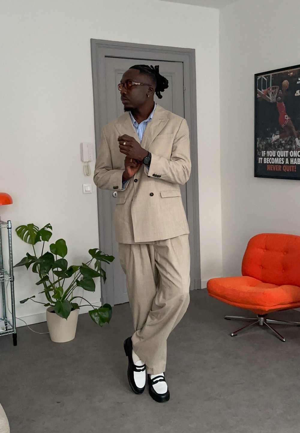 Homme en costume beige et chaussures blanches debout à l'intérieur près d'une porte grise, une plante d'intérieur, une chaise orange et une affiche de basketball sur le mur.