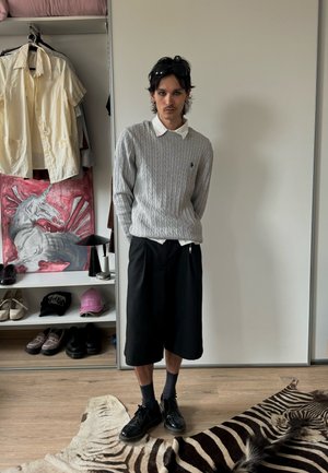 Ung man med kort mörkt hår, klädd i grå tröja, vit skjorta, svarta shorts och svarta skor, står inomhus nära en garderob och ett zebrarandigt matta.