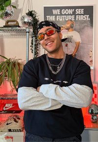 Homme souriant portant un bandana et des lunettes de soleil, croisant les bras devant des étagères avec des plantes, des chaussures, des boules disco, et une affiche "We gon’ see the future first".