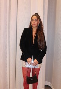 Čierny sametový blazer, metalická strieborná mini sukňa, červené čipkované silonky, malá čierna kabelka. Dlhé, vlnité vlasy a nápadné náušnice dopĺňajú vzhľad.