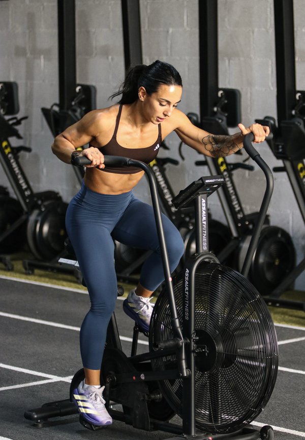 Femme en forme en tenue de sport avec des pédales tatouées et des poignées sur un vélo ventilateur noir dans une salle de sport avec des machines d'exercice en arrière-plan.