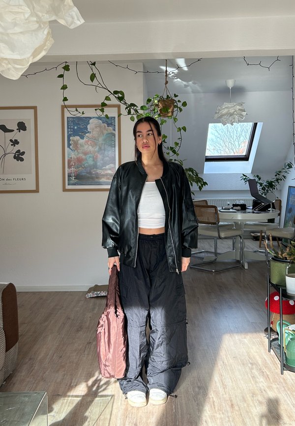 Jeune femme debout à l'intérieur, portant une veste en cuir noire, un top blanc court, un pantalon large noir, tenant un sac rose, avec des plantes et des œuvres d'art encadrées derrière elle.