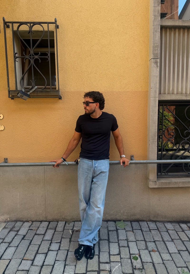 Homme portant une chemise noire et des lunettes de soleil, appuyé sur une rambarde en métal contre un mur jaune avec des fenêtres grillagées dans une rue pavée.