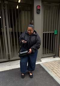 Giacca nera imbottita; camicia nera; pantaloni in denim a gamba larga; borsa a tracolla nera; scarpe piatte e texturizzate; in piedi vicino a un ingresso con cancello.