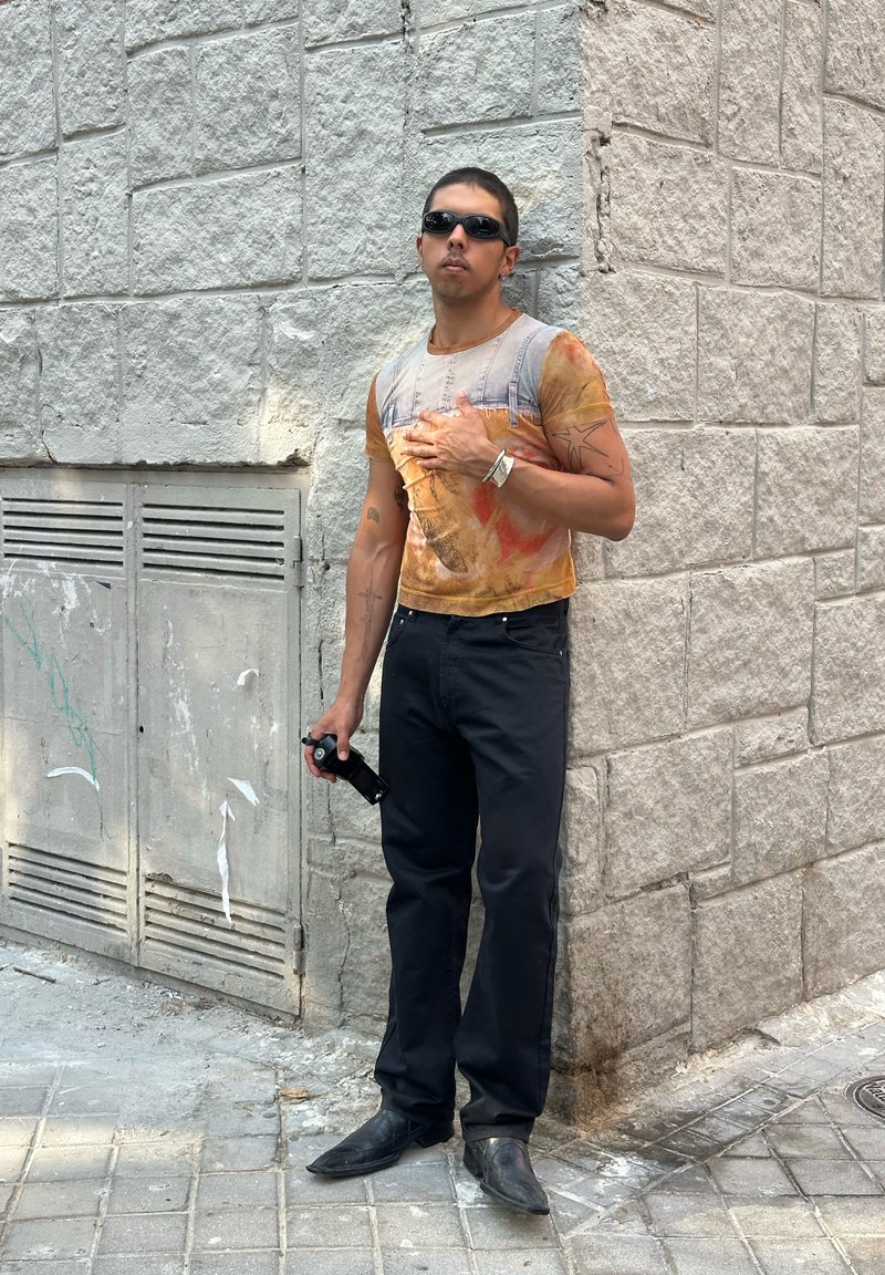 Άνδρας με γυαλιά ηλίου, πολύχρωμο κοντομάνικο πουκάμισο, μαύρο παντελόνι και μπότες στέκεται μπροστά σε έναν πέτρινο τοίχο κρατώντας ένα μαύρο αντικείμενο.