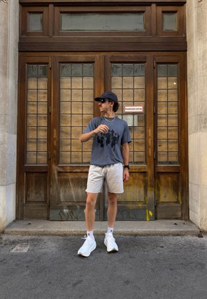Joven con gafas de sol, gorra, camiseta oscura, pantalones cortos claros y zapatillas blancas, de pie delante de grandes puertas de madera con paneles de cristal.