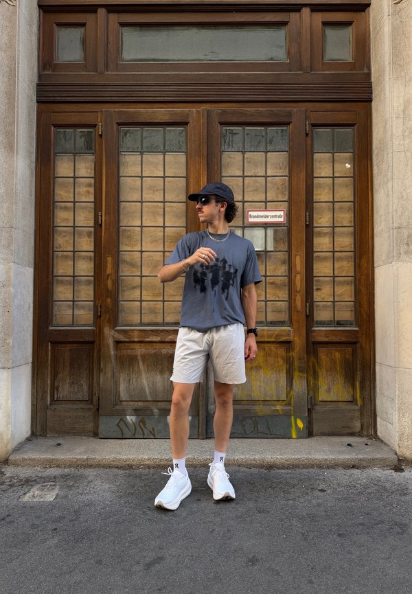 Junger Mann mit Sonnenbrille, Mütze, dunklem T-Shirt, hellen Shorts und weißen Sneakers steht vor grossen Holztüren mit Glaseinsätzen.