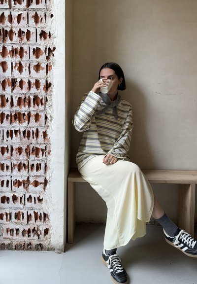 Mujer con cabello corto y oscuro sentada en un banco de madera junto a una pared de ladrillos ventilada, vestida con un suéter de rayas, falda crema, calcetas grises y zapatillas negras, bebiendo de una taza.