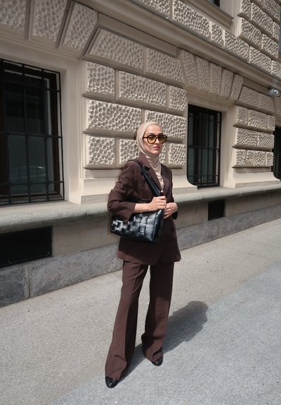 Traje marrón a medida con pantalones de pierna ancha, combinado con un hijab beige y gafas de sol amarillas de gran tamaño. Un bolso negro de mimbre completa el conjunto.